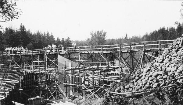 Two groups of men are standing on a temporary wooden bridge over the Totogatic River. There is a large form in place on the left and a complete concrete wing wall, center. On the reverse of the photograph is written: "Construction work on the Lake Nancy dam on the Totogatic River." The Totogatic River is a tributary of the Namekagon River.