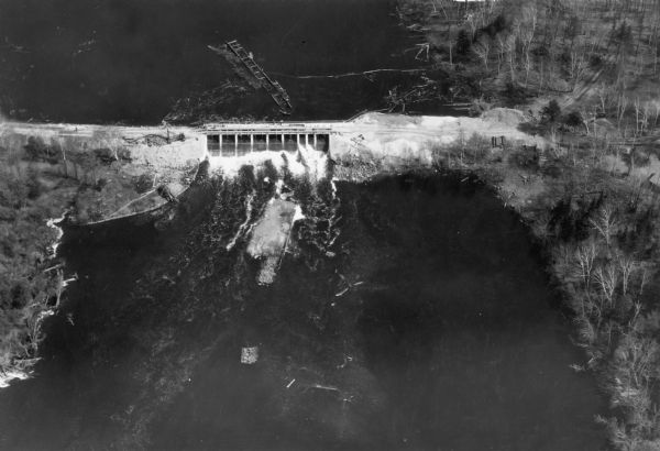 Aerial view, looking upstream, of water flowing through a dam. On the reverse of the print is written: "Dam built by WPA on site of old logging dam on the St. Croix near Gordon. A new lake more than seven miles in length will take the place of the one which disappeared when the old dam washed out." There are logs in the river above the dam and construction equipment and materials on the shore.