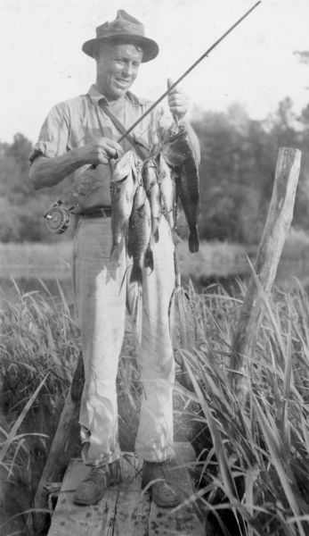 Jean A. Pope is smiling broadly while holding a stringer of five bass. He is also holding his rod and reel, and is standing on a small wooden pier. A description on the reverse of the photograph states that the bass were caught in the St. Croix River, which is in the background.