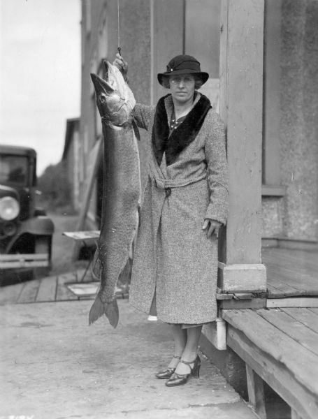 A well-dressed, unidentified woman is standing next to a muskie hanging from a line. She is next to the porch of a large building, and there is an automobile in the background. The description on the reverse of the photograph reads: "Muskie, 50" long, weighed 43½ pounds, just about 10 pounds lighter than his heaviest relative caught in Wisconsin in 1935. Caught in Little Papoose Lake."