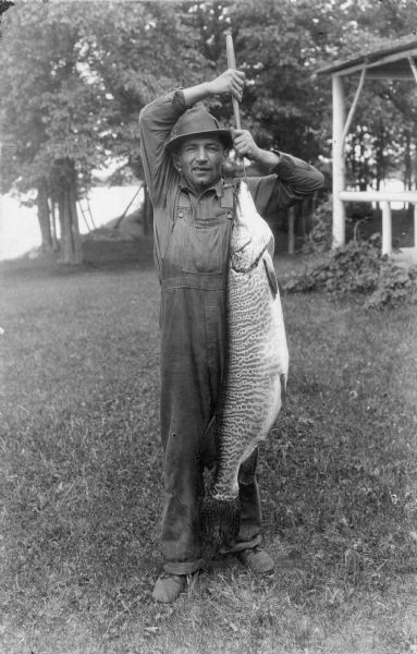 An unidentified man standing on a lawn is holding a 48 pound muskie by a wood handled hook. There is a lake in the background, and on the right, the rustic framing and rail of a porch. The man is wearing bib overalls and a hat.
