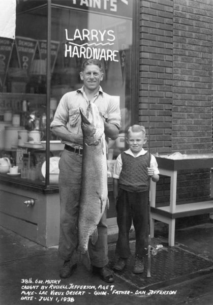 Seven-year-old Russell Jefferson, right, is standing holding his rod and reel next to his father, Dan Jefferson. Dan is holding the 38 pound, 6 ounce muskie which Russell caught at Lac Vieux Desert. Both father and son are beaming as they are standing in front of Larry's Hardware store. Banners in the store are advertising "Demonstration Days." There is crockery and enamelware displayed in the window. Caption reads: "38 lb. 6 oz. Musky, Caught by Russell Jefferson, Age 7, Place -- Lac Vieux Desert - Guide -- Father -- Dan Jefferson, Date -- July 1, 1938."