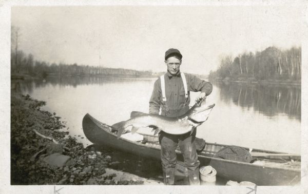 An unidentified man wearing waders and a cap is posing on a rocky shore holding up a large muskie. There is a wooden canoe in the shallow water behind him.
