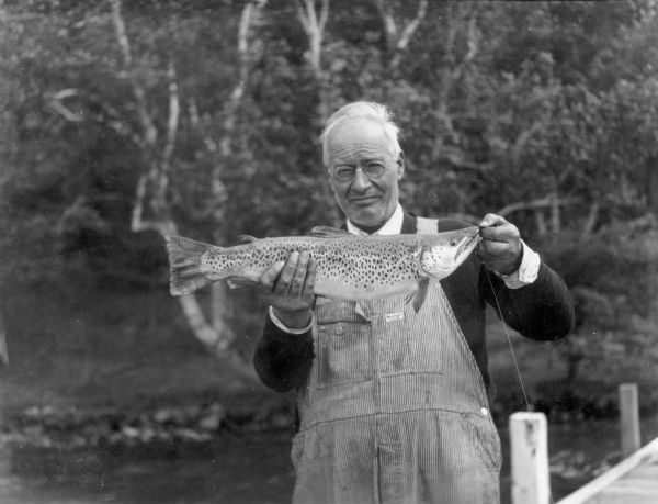 An unidentified man wearing bib overalls is posing on a pier holding a large trout. On the reverse of the photograph is written: "An 8½ lb. trout taken from Wisconsin waters."