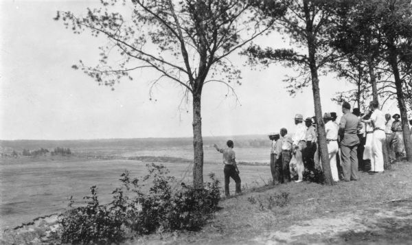 A crowd has gathered on a hilltop overlooking a large flat expanse which has been cleared of trees. A man is standing slightly downhill from the group and is pointing over the area. A description on the reverse of the photograph reads: "Eau Claire County citizens view the cleared flowage area of the future Lake Eau Claire, located within their county forest."