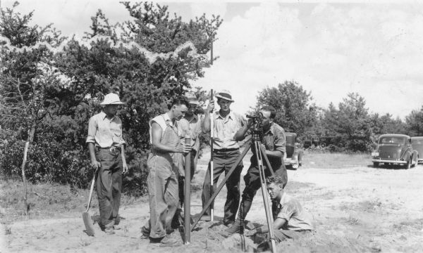 One man is sighting a transit level, and another man is holding a round metal post as a crew of six people "sets a corner" in a Jackson County forest. There are automobiles in the background.