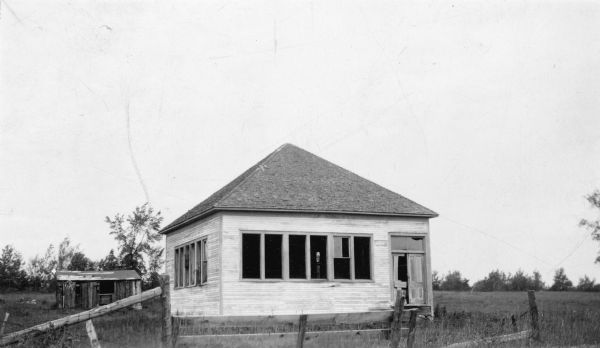 A simple one-room schoolhouse with a hip roof is standing behind a neglected fence. Most of the window glass is gone and the paint has faded. There is a derelict outbuilding in the background. A description on the reverse of the photograph reads: "Two conditions are principally responsible for abandoned school: the settlement on lands unfit for agriculture and the loss of the forests which once furnished winter employment for settlers."