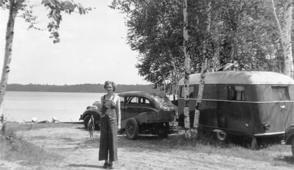 A young woman is standing and posing with a stringer of panfish in front of a car with a small travel trailer behind it. She is wearing a wide leg bib overall style playsuit. There is a two wheeled wagon near the car. Birch trees shade the campsite which is on the shore of Carrol Lake in the Northern Highland American Legion State Forest.
