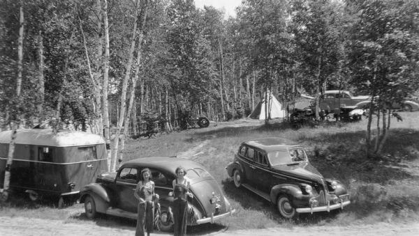 Elevated view of two young women, wearing nearly identical bib overall-style playsuits, are standing posing holding stringers of pan fish. There are three automobiles in the background. There is a teepee in the background, and a travel trailer in the foreground on the left. The campsites, located on the shore of Carrol Lake in the Northern Highland American Legion State Forest, are shaded by birch trees.