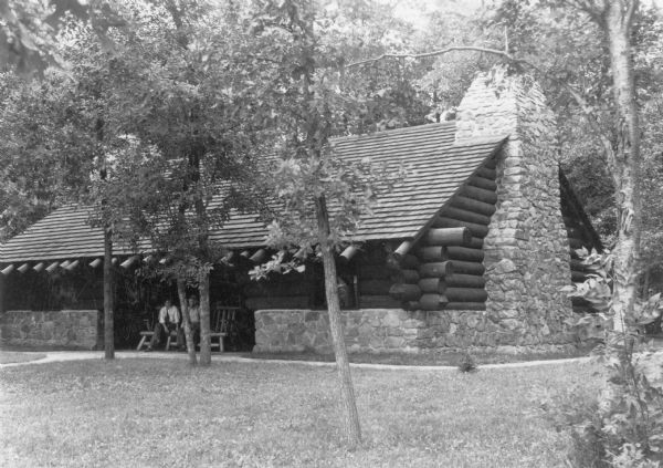 View across lawn towards two men sitting on a log bench in the central open area of the shelter house at Council Grounds State Forest (now Council Grounds State Park). The shelter is built of sturdy logs, with a massive stone chimney on the right. The stonework continues as a half wall at the base of the building. 