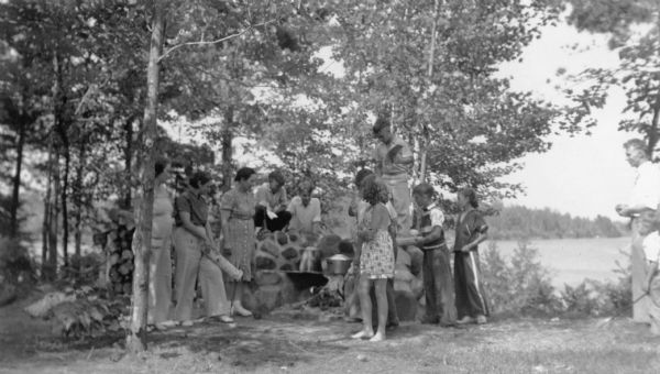 Three unidentified women, two wearing wide-legged slacks, are supervising a group of children around a stone fireplace in Council Grounds State Forest (now Council Grounds State Park). One woman is holding a piece of firewood. There is a large coffee pot and covered kettle on the grate. A man and boy are standing at far right. Lake Alexander is in the background.