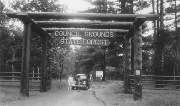 Two well-dressed women are posing next to an automobile parked near the arbor-like gate at the entrance to the Council Grounds State Forest, now the Council Grounds State Park. The gate is constructed from massive logs, and there is a log rail fence on either side of the entrance. "USA WPA WORKS" is printed on the sign in the background.  