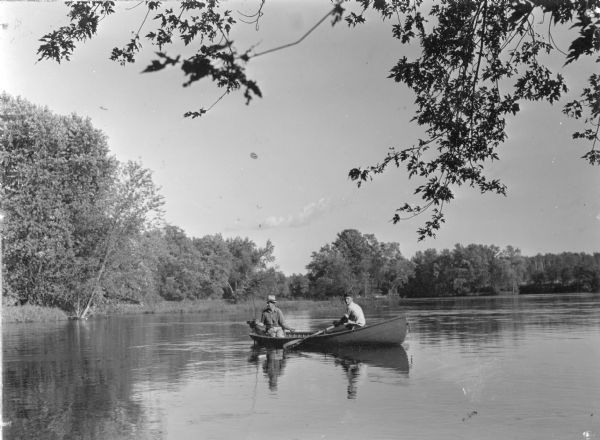 View across water towards an unidentified man sitting in the stern of a small boat, holding a fishing rod. A younger man, sitting forward, is rowing. There is a small trolling motor on the boat. On the reverse of the photograph is written: "Boating on the Wisconsin River near Council Grounds State Forest [now Council Grounds State Park]".    