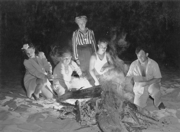 A group of people, including two men, two women and a youth, is gathered around a campfire on a beach in the Kettle Moraine State Forest. One of the women, who appears to be older, is standing behind the others as they toast marshmallows. The woman at far left has a flower in her hair. A written caption on the reverse of the photograph reads: "Picnic fire in the Greenbush Sports Area."  