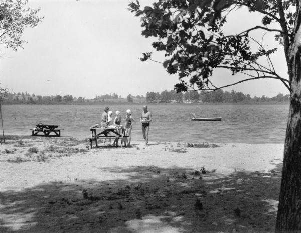 View across beach towards a woman wearing a dress sitting on the top of a picnic table. Three girls in swimming suits and caps are standing near her. A lifeguard, identified by "Guard" printed on his top, is standing on the right. In the background there are two girls sitting on a wooden raft in the lake. 