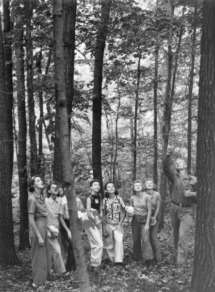 An unidentified man is leading a group of young women on an educational walk in the Kettle Moraine State Forest. He is pointing upward near a large tree. The women are wearing wide legged slacks and summer blouses. The two women at right appear to be twins.