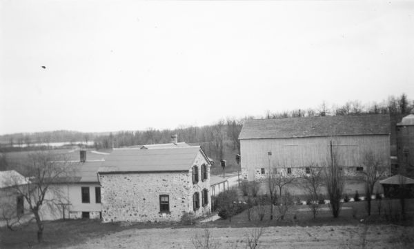 An elevated view of a farmstead, including a house, barn, silo, and two small outbuildings. The house has a fieldstone wing with front gable and shutters on the four front windows; a rear wood framed addition and the roof of another wing with porch are visible. The large barn has a fieldstone foundation. There are two birdhouses mounted on tall posts between the house and the barn. The buildings were used as the headquarters of the Kettle Moraine State Forest, Northern Unit. Mauthe Lake is visible in the background, left.