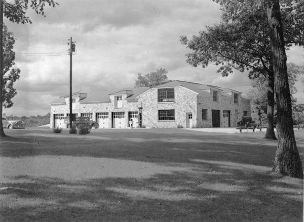 Trucks and an automobile are parked near a large stone clad building with multiple garage bays and large windows. There is a gasoline pump near the building. A caption on the reverse of the photograph reads: "Garage and work shop at Mauthe Lake, Kettle Moraine State Forest." 