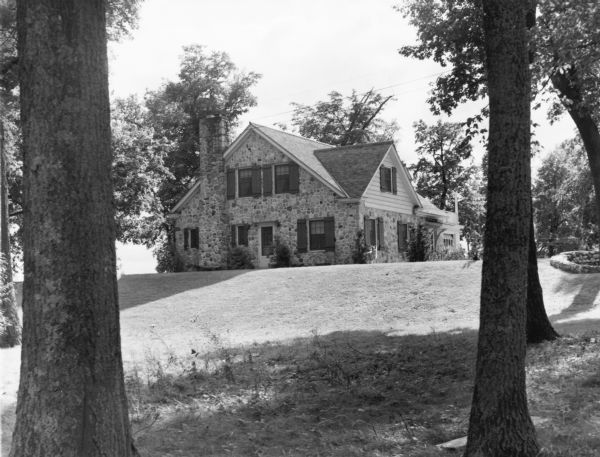 A one and one half story, stone clad house with wide siding in the front gable is standing on a low rise surrounded by a lawn and mature trees. On the reverse of the photograph is written" "Ranger home at Kettle Moraine State Forest."  
