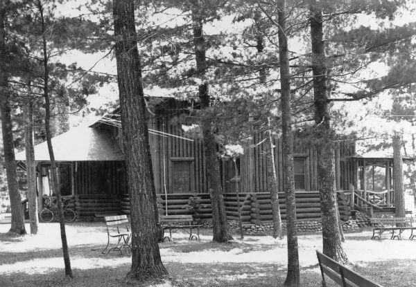 A log house with a cobblestone foundation stands among pine trees in the Northern Highland State Forest. Horizontal logs comprise the lower portion of the walls; the upper portion is made of vertical logs. There are rustic front (right) and back porches. A bicycle is leaning against the back porch. There are park benches on the lawn. On the reverse of the photograph is written: "Home of the Chief Forest Ranger, Trout Lake, Wisconsin."