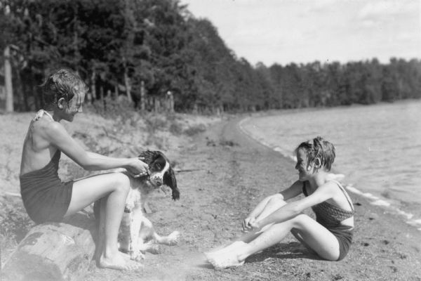 The caption on the reverse of this photograph reads: "Steiro girls sitting on lake shore at Trout Lake." The girls are wearing swimming suits, and a dog is sitting on the sand next to the girl at left. She is holding a stick which the dog has clenched in its mouth. Trees line the curving shoreline.