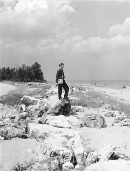 An unidentified man is standing on a boulder on the beach looking out at Lake Michigan.  