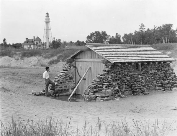 A man is standing and looking at the door of the boathouse on the beach near the Twin River (Rawley) Point Lighthouse. The boathouse has stone sides and a log front. A log is propped against the door, which has a massive wrought iron hinge. The lighthouse and keeper's house are in the background.  