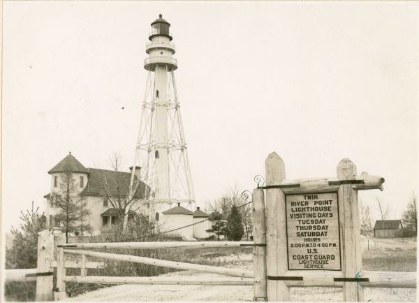 A log gate and log-framed sign mark the entrance to the Twin River (Rawley) Point lighthouse. The keeper's house is next to the light and has a turret and porch. There are three low structures with roof ventilators near the light.