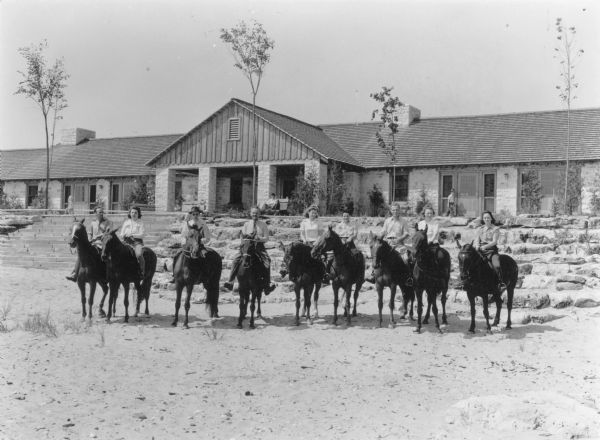 Nine unidentified men and women on horseback are posing in front of the lodge at Point Beach State Forest. A young girl wearing a swimming suit is sitting on a railing in the background on the left, and a young man is sitting on a bench in the center. Another boy is walking on the right. Young trees are standing in front of the long one story building. which has two massive stone chimneys, and a wide stone stairway that leads down to the beach. A gabled porch extends from the center of the building.
