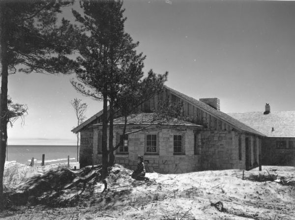 An unidentified woman is posing sitting on the sand and looking toward Lake Michigan. The newly built shelter house, with its large windowed bay and massive chimney is behind her. There are pine trees in the foreground.  