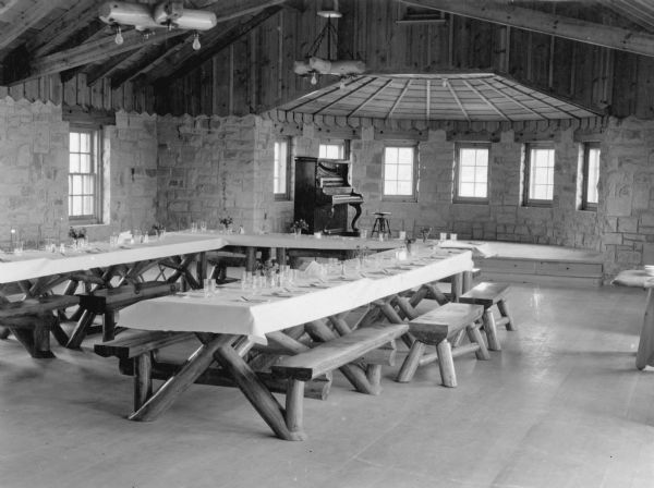 A view of the dining room in the shelter house at Point Beach State Forest. There are sturdy log picnic tables set with glasses, silverware and tablecloths. The bench seats are single split logs. There is a bay at the end of the room with a raised stage and piano. The walls are bare stone, and there are rustic log chandeliers overhead.