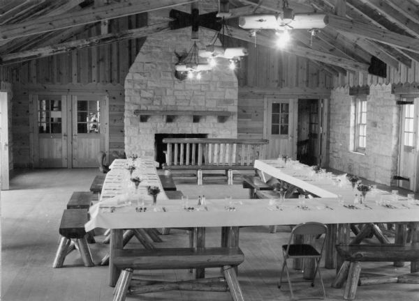 A view of the dining room at Point Beach State Forest. Tables are arranged in a "U" formation and are set with silverware, glasses and tablecloths. The rustic log benches have seats made from a single split log. There are log chandeliers overhead and a large fireplace with mantel. A mounted deer head is hung on the chimney. The exterior walls are exposed stone. The timber frame rafters with king posts are also exposed.