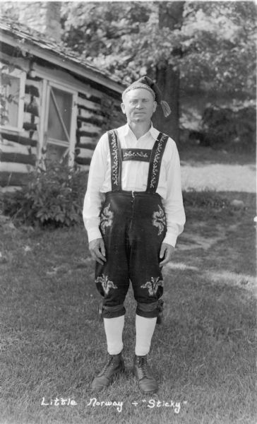 A man identified only as "Sticky" is posing standing on the lawn in front of a log cabin at Little Norway. He is wearing traditional Norwegian dress with embroidery on his suspenders and leggings.