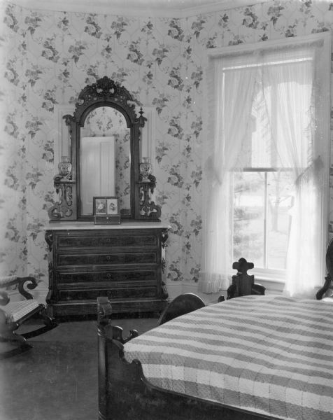 A view of the southwest bedroom of Villa Louis, the Hercules Louis Dousman mansion. There is floral wallpaper on the walls, and the room is furnished with a large dresser with mirror, a rocking chair and bed. There are sheer curtains on the window and a coverlet on the bed.
