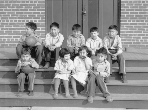 A group of nine children, two girls and seven boys, sitting on the steps of the Indian School at Tomah. Two boys on the left are holding balls. All of the children are wearing stockings but no shoes. The girls are wearing light-colored dresses, and the boys have matching side stripe pants, and various styles and colors of shirts.  