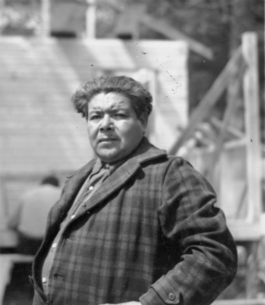 Waist-up outdoor portrait of a Native American man. He is wearing a plaid jacket over a dark-colored shirt. There appears to be construction in progress behind him. The description on the reverse of this print reads: "George W. Brown, president of the Tribal Council, Lac du Flambeau Band of Lake Superior Chippewa Indians."