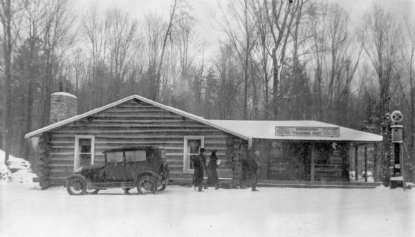 Two men and a woman are walking from a parked automobile toward the covered porch of the Oshkosh Trading Post, a low, log building on the Menominee Indian Reservation. There is a Sinclair HC gasoline pump on the right. A sign over the porch advertises Indian baskets, bead work, novelties, ice cold drinks, cigarettes and candy. There is a stone chimney on the left. It is snowing and there are a few inches of snow on the ground.  