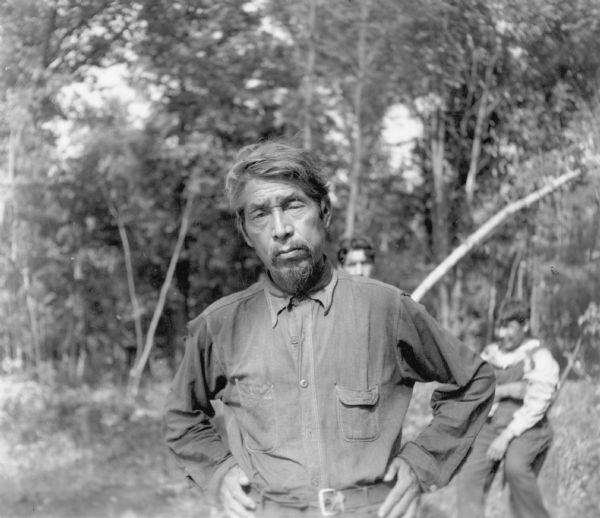 A waist-up informal portrait of Match-o-ka-mah, described on the reverse of the photograph as "Medicine Man and Keeper of the Sacred Drum of the Wisconsin Menominee Indians." He is wearing a work shirt and has a small beard and mustache. There are two unidentified persons in the background.