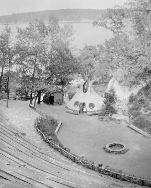 An elevated view towards a Native American man wearing a feathered headdress and a young girl of European descent standing in front of a tepee on the stage area of the ceremonial grounds at Stand Rock, and just behind are two wigwams and a second tepee. There is a stone fire ring on the stage on the right. Bleachers are on the slope of the hill in the foreground. The Wisconsin River and the far shoreline is in the background.