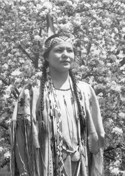 Waist-up outdoor portrait of Cecelia Blackdeer (1917-1990), the daughter of Charles and Carolind Decorah Blackdeer standing in front of a flowering tree. She is wearing a traditional Ho Chunk deerskin dress with embroidery over the shoulders.