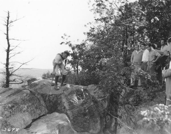 A male guide on the left is using a walking stick to point out a feature in the rocks atop a bluff at Devil's Lake State Park, as a group of men on the right are looking on. The guide is wearing knee-high lace-up boots.