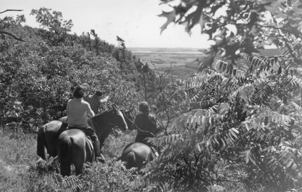 Two women and a man (a fourth rider is hidden) on horseback are enjoying the view from a bluff in Devil's Lake State Park. The Devil's Doorway rock formation is in the distance. The tip of Devil's Nose, the rounded terminus of a bluff, is in the background on the right. Lake Wisconsin is in the distance. There is a stand of sumac in the right foreground.