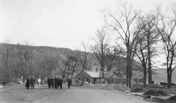 Men wearing hats and long overcoats are walking toward the camera from the bathhouse at Devil's Lake State Park. Other men in work clothes, members of the Civilian Conservation Corps (CCC), are working on the building. Two men are on the roof; others are working on the ground shoveling and sifting piles of aggregate. There are no leaves on the trees. The East Bluff is in the background and there is a glimpse of the lake on the right. On the reverse of the print is written: "State Foresters inspecting the new bath house [sic] under construction at Devil's Lake."