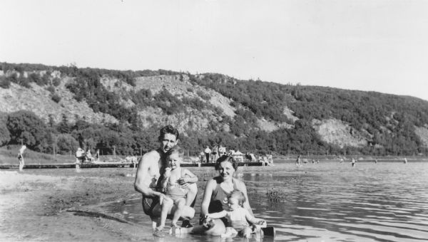 A man and his wife are wearing swimming suits and posing sitting on the beach. Each of them is holding a small child in their laps. People are standing and sitting on a long pier behind them. The East Bluff is in the background. On the reverse of the print is written: "Enjoying the beach at Devil's Lake State Park. Mr. & Mrs. F.E. Wedel and family, 524 Second St., Baraboo, Wis."