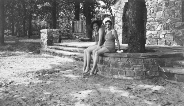 Two women in bathing suits and wearing brimmed hats are posing sitting on a stone wall. The description on the reverse of the print reads: "Two fair bathers posing for a picture i n front of the new bathhouse at Devil's Lake State Park."  