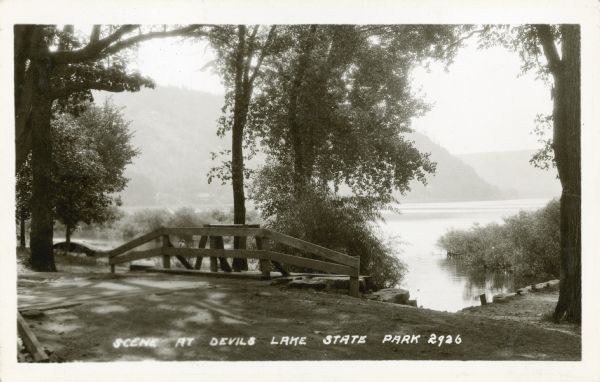 The bluffs and lake appear hazy beyond an unpaved road and simple wooden bridge at Devil's Lake State Park. A man or boy at the shoreline can be seen through the bridge's railing. There is a canoe on the beach on the left. Caption reads: "Scene at Devil's Lake State Park."