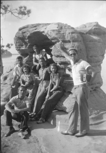 A young man wearing a cap and sunglasses is posing standing with his right hand resting on a rock formation. He is holding a Kodak Brownie No. 2A box camera. Six other young people, three men and three women, are posing on the left in and around a large opening in the rock. The women are wearing slacks. Devil's Lake is in the background. A caption on the reverse of the print reads: "Hikers resting at one of the picturesque rock formations at Devil's Lake State Park."