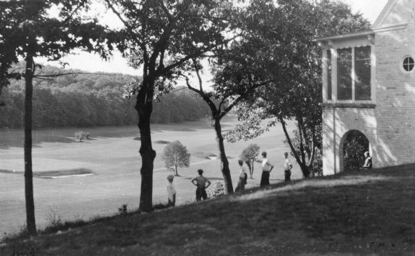 View down hill towards six men enjoying an elevated view of the golf course at Devil's Lake State Park. One of the men is sitting under the porch of the brick clubhouse on the right.
