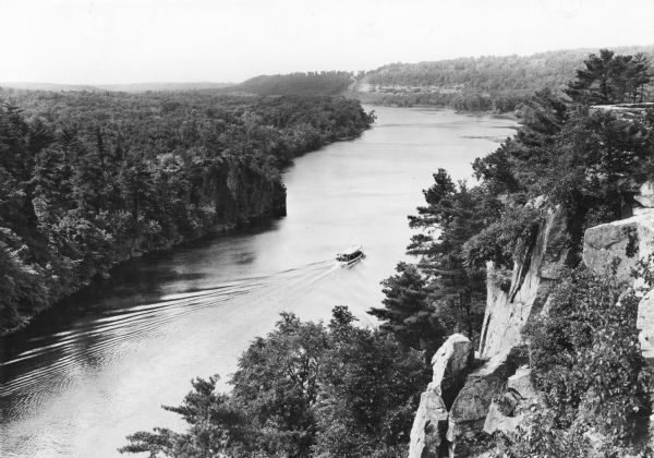 An elevated view of the St. Croix River at Interstate Park. There is a boat with open sides, probably an excursion boat, going down the river, and rock formations are on the right. The stone wall of an observation area is on the right at the top of the bluff.  