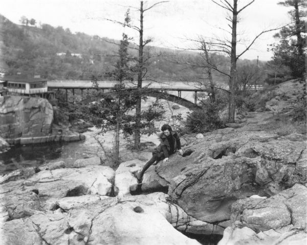 A woman identified as Beth Doane is sitting on a rock formation high above the St. Croix River. She is looking into one of several potholes in the area. The Show Boat Inn of Taylor Falls, Minnesota is on the left. The bridge in the background, at the northern end of the Dalles of the St. Croix, connects Wisconsin with Minnesota.